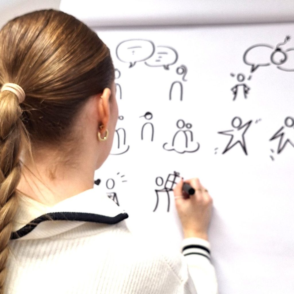 Frau mit Zopf zeichnet einfache Figuren auf ein Flipchart.